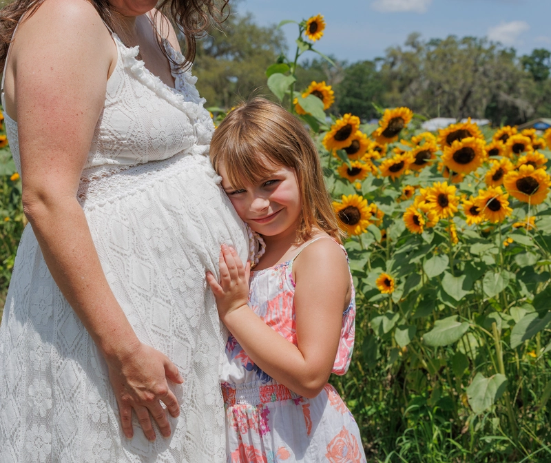 A pregnant woman in a white dress stands in a field of sunflowers with a young girl in a floral dress gently touching her belly.