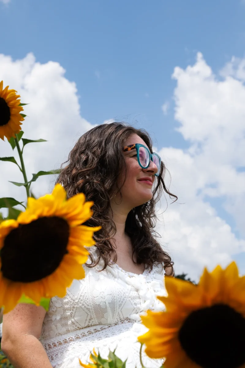 A woman stands amidst vibrant sunflowers, with a bright blue sky and fluffy clouds in the background, radiating summer joy