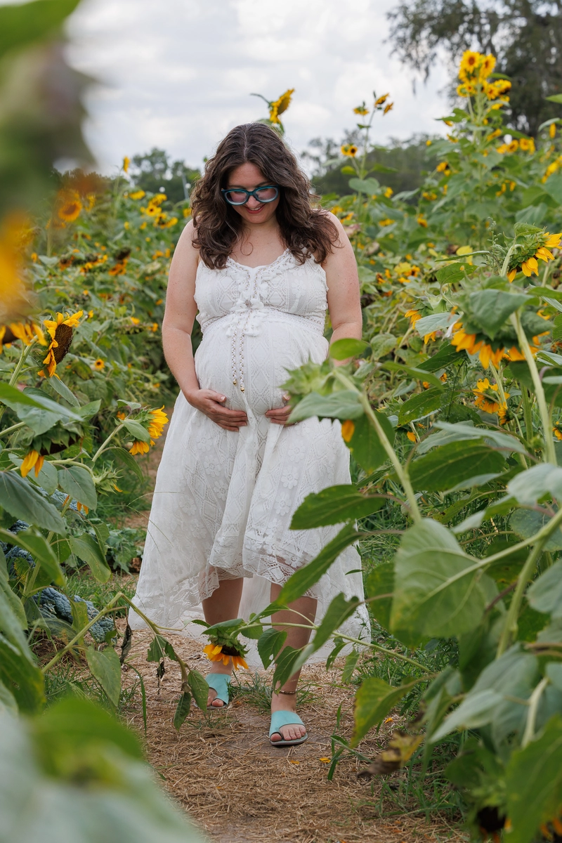 A woman in a flowing white dress stands in a sunflower field, gently cradling her pregnant belly. Sunflowers surround her, creating a vibrant scene.