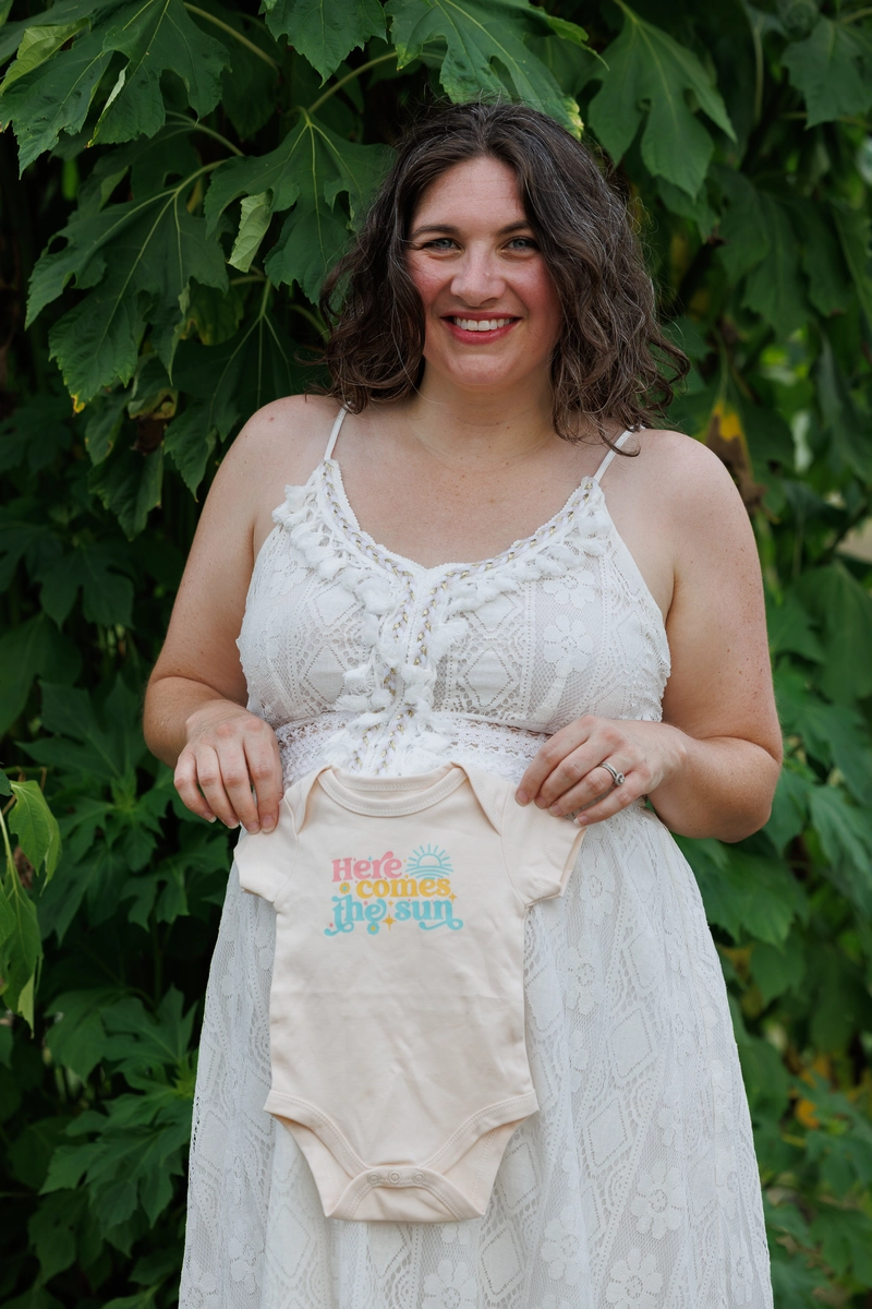 A woman in a white dress holds a beige onesie with the text "Here comes the sun," surrounded by lush green foliage.