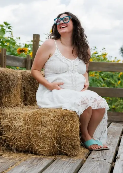 A pregnant woman in a white lace dress sits on hay bales, surrounded by green foliage and sunflower blooms under a cloudy sky.